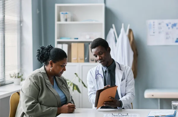 Portrait of young black doctor consulting woman in clinic and pointing at tablet screen, copy space
