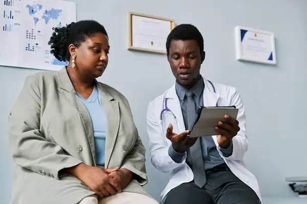 Low angle portrait of young black doctor and patient talking in clinic and looking at tablet screen