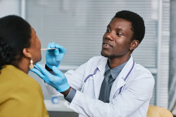 Portrait of young black doctor examining patient in clinic and doing flu check up