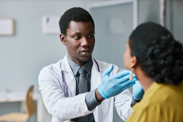 Portrait of young black doctor consulting patient in clinic and doing throat exam with palpation