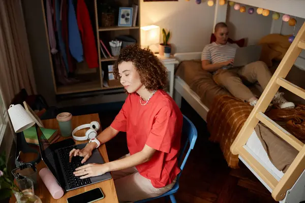 High angle view at two female students in dorm room with smiling young woman using laptop in foreground, copy space