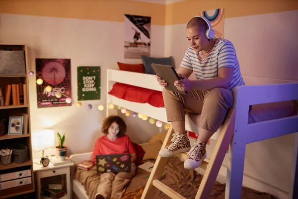 Portrait of students dorm room with bunk bed and two young women using tech devices, copy space