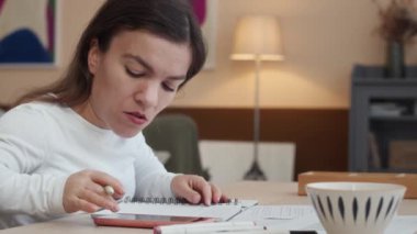 Beyaz tenli, kısa boylu, evrak işleriyle uğraşan, hesap makinesi kullanan, oturma odasında masa başında oturan genç bir kadının belden yukarı görüntüsü.