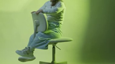 Kısa boylu neşeli bir kadının yavaş yavaş portresini kaldır ofis sandalyesinde dön ve neon yeşil stüdyo arka planındaki kameraya gülümse.
