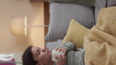 Modern oturma odasındaki rahat koltukta oturan ve dizüstü bilgisayarında bir fincan çay içip komedi filmi izleyen genç, beyaz bir kadının dikey çekimi.