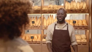 Önlüklü, genç, Afro-Amerikan erkek fırın işçisinin omzunun üstünde taze ekmekle rafların karşısında durup beyaz kadın müşteriyle konuşuyor.