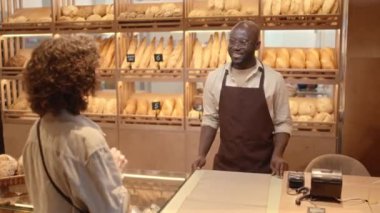 Orta boy siyah erkek satıcının tezgahta durup, beyaz kadın müşteriyle konuşurken, küçük bir fırında çalışırken taze ekmek ve hamur işi satarken.