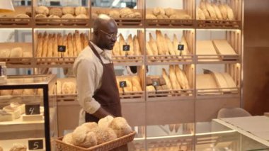 Önlüklü, genç, kel, erkek fırın işçisinin orta yavaş portresi. Tezgahta duruyor ve kameraya gülümsüyor, taze fırınlanmış mallar ve tatlılar satıyor.