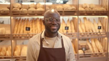 Önlüklü ve gözlüklü genç Afro-Amerikan kel satıcının belden aşağı portresi raflarda duran kameraya gülümsüyor taze ekmek ve baget ekmeğiyle