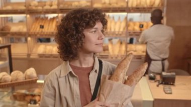 Beyaz saçlı, kıvırcık saçlı, kağıt torbada iki uzun taze baget tutan ve rahat fırında dururken kameraya gülümseyen genç kadının yavaş yavaş belini kaldır.