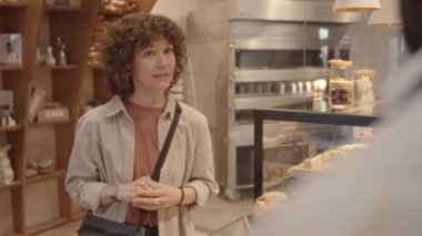 Omzunun üstünden, kıvırcık saçlı beyaz bir kadının tezgahtaki fırın işçisine sipariş verirken orta boy bir fotoğrafı.