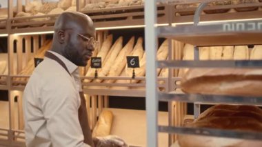 Kahverengi önlük ve tek kullanımlık eldivenlerle sabahları tepsilerden raflara taze ekmekler sererken orta boy zenci fırın işçisinin fotoğrafı.