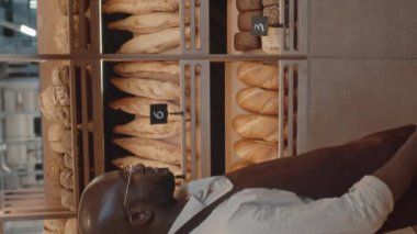 Önlüklü genç Afrikalı Amerikalı fırın satıcısının dikey yavaş portresi katlanmış ellerle taze baget ve kameraya gülümsüyor.