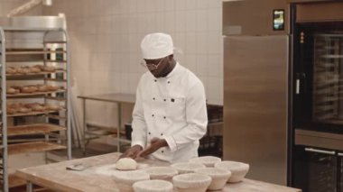 Orta boy Afro-Amerikan erkek fırıncı beyaz üniformalı ahşap çalışma masasında durup hamur yoğururken fırında ekmek yaparken.