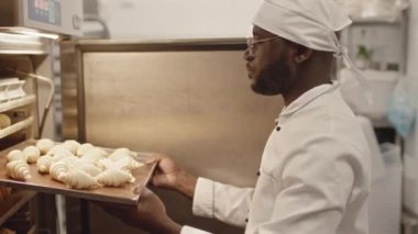 Beyaz üniformalı ve gözlüklü genç siyahi aşçının beline fırında çiğ kruvasanla pişirme tepsisi yerleştirirken pastanede çalışıyor.