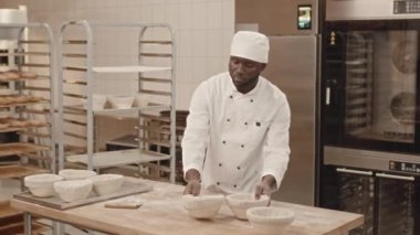 Orta boy Afrika kökenli Amerikalı fırıncı, fırın mutfağında çalışırken yükselmesi için rafa bir kase çiğ ekmek hamuru koyuyor.