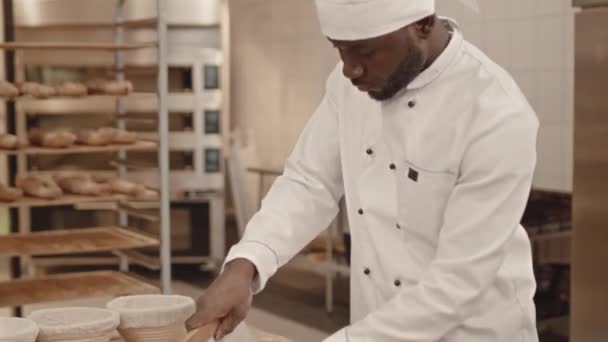 ベンチスクレーパーを使用した濃縮された若い黒人男性ベーカのミディアムショットは パンとペストリーをパン屋の木製テーブルに作りながら生地をカットします — ストック動画