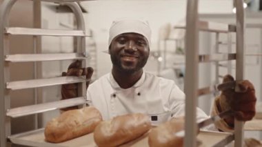 Genç Afrikalı Amerikalı fırın işçisinin belden yukarı portresi kameraya poz veriyor fırının mutfağındaki alüminyum tepside taze ekmeklerle.