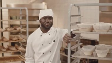 Orta boy gülümseyen siyah erkek fırıncı üniforması içinde tepside ekmek sepetleriyle eğilmiş ve fırın mutfağındaki kameraya poz veriyor.