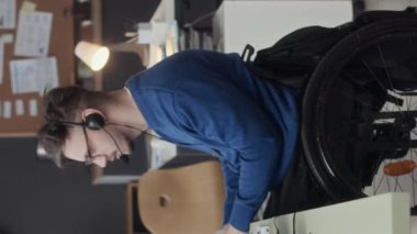 Tekerlekli sandalyedeki genç beyaz adamın mikrofonlu kulaklık taktığı ve çağrı merkezinde çalışırken müşteriyle konuştuğu orta boy bir fotoğraf.