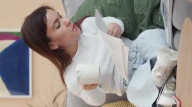 Modern oturma odasındaki koltukta oturan, mali raporlara ve belgelere bakan ve evde çalışırken dizüstü bilgisayar kullanan genç iş kadınının dikey görüntüsü.