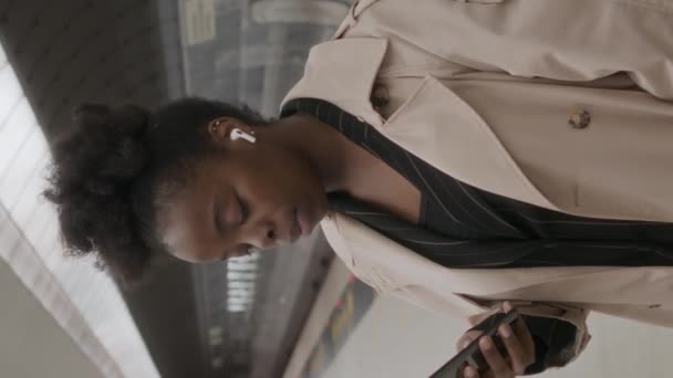 坐在无线耳机里的年轻黑人妇女站在地铁站旁 环顾四周 一边等火车一边用智能手机 拍下了一个垂直的中等照像 — 图库视频影像