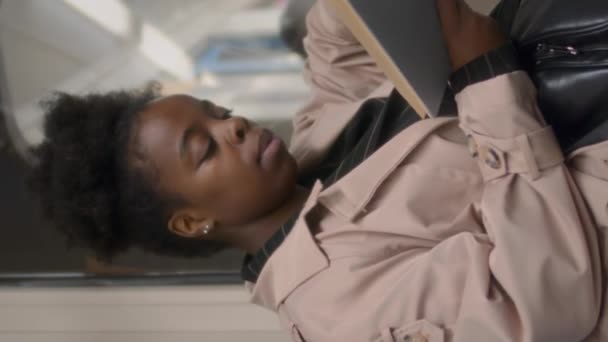 年轻黑人女子独自坐在地铁窗口看书时的垂直中拍 — 图库视频影像