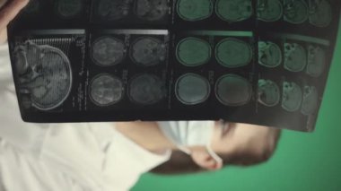 Beyaz laboratuvar önlüğü ve yüz maskesi takmış beyaz bir kadın doktorun dikey portresi hastanın beyin röntgenini çekiyor sonra da yeşil stüdyo arka planında duran kameraya bakıyor.