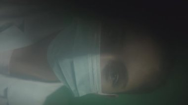 Mavi gözlü beyaz kadın doktorun dikey portresi. Yüz maskesi takmış hastalara bakıyor. Röntgen ve kamera, açık yeşil arka planda duruyor.