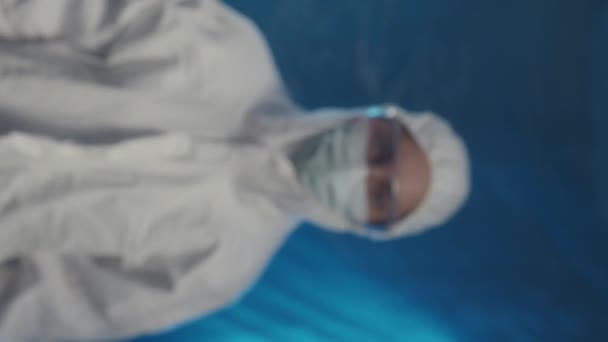 Vertical chest up portrait of Caucasian female doctor wearing safety coverall, glasses and face mask looking at camera standing on blue background