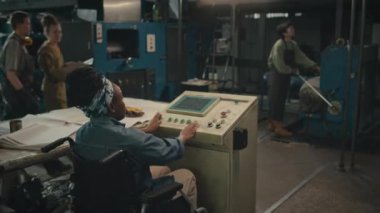 Arka planda, diğer çeşitli kadınlarla fabrikada çalışırken mavi kot gömlek ve bandana takan genç siyahi bir kadın görülüyor.
