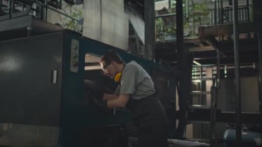 Endüstriyel fabrikada diğer kadınlarla çalışırken omuzlarında koruyucu kulaklıkları olan orta boylu, beyaz bir kadının makine ekipmanına baktığı bir fotoğraf.