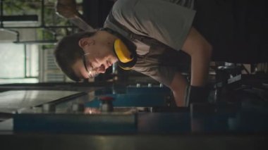 Endüstriyel fabrikadaki makine ekipmanlarını tamir ederken güvenlik gözlüğü ve kulaklıklı genç beyaz kadın tamircinin dikey görüntüsü.