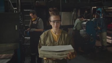 Kameraya poz veren, güvenlik gözlüklü beyaz bir kadının orta boy portresi. Elinde yeni üretilen kağıt yığınıyla matbaada çalışan diğer kadınlarla birlikte.