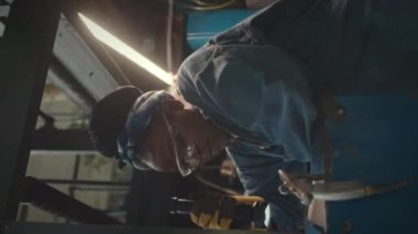 Endüstriyel fabrikada makine ekipmanlarını tamir ederken el feneri kullanan Afro-Amerikalı bir kadının güvenlik önlüğü ve mavi kot gömlek giydiği dikey bir fotoğraf.