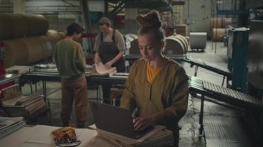 Baskı fabrikasında diğer kadınlarla çalışırken genç beyaz bir kadının dizüstü bilgisayar kullandığı orta boy bir fotoğraf.