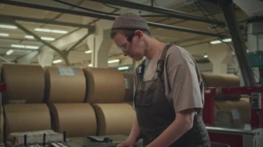 Baskı fabrikasında üretim hattının yanında duran ve üretim kağıdının kalitesini kontrol eden güvenlik gözlüklü kadın işçinin orta boy fotoğrafı.