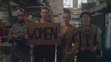 Feminist sembolleri ve sloganları olan mukavva pankartlar taşıyan ve endüstriyel fabrikada kamera önünde poz veren genç kadın aktivistlerin orta yavaş portresi.