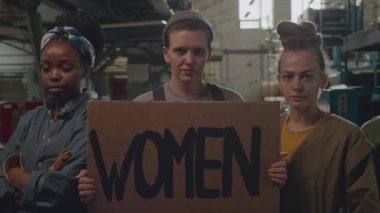 Feminist sembolleri ve karton pankartlarda yazılı sloganları olan ve eski endüstri fabrikasındaki iş yerinde kameraya poz veren genç aktif kadınlardan oluşan PAN toplumunun orta derecede yavaşlaması