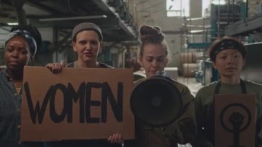 Feminist afişlerle ve sözcülükle sloganlar atarak endüstriyel fabrikada kameraya poz veren dört etkin kadının orta karar görüntüsü.