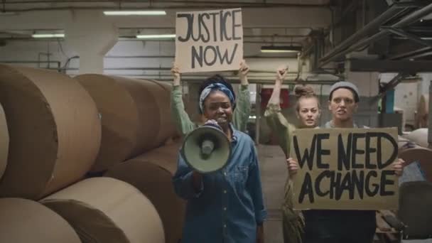 マウスピースで話す若い黒人女性と 産業工場で集会しながら彼女をサポートする抗議バナーを持つ他の多様な女性のミディアムショット — ストック動画