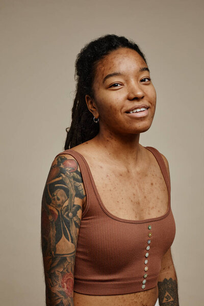 Vertical portrait of young black woman looking at camera standing against neutral beige background with focus on tattoo and real skin texture