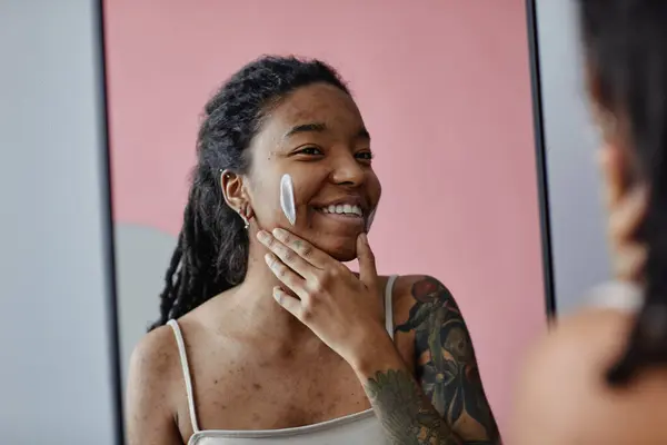 Candid portrait of smiling black woman with acne scars using face cream by mirror