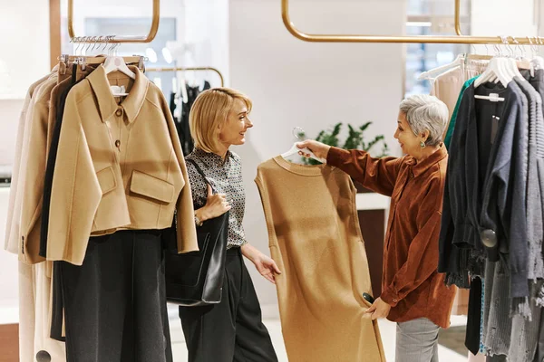 Side view portrait of sales assistant helping senior woman choosing clothes in luxury boutique