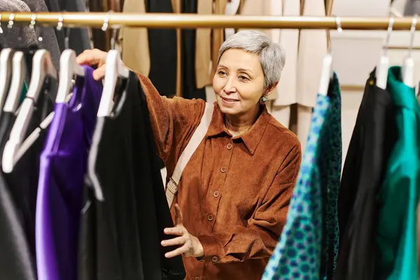 Front view of smiling senior woman browsing clothes on racks in luxury boutique, copy space