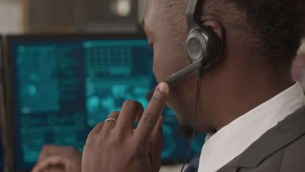アフリカ系アメリカ人の男性サイバーセキュリティ担当者が 秘密のネットワークデータプログラムでコンピュータモニターを見ながら 1つの耳のマイクヘッドセットに話しかける中間閉鎖 — ストック動画