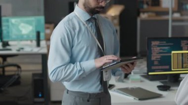 Modern siber güvenlik bürosunda dikilirken elinde dijital tabletle kameraya bakan sakallı, çift ırklı erkek siber güvenlik görevlisinin orta yavaş portresi.