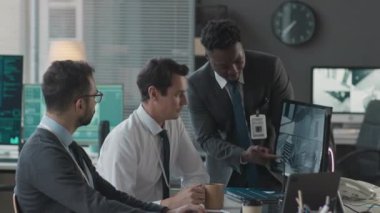Çoklu CCTV kameralarıyla bilgisayar ekranına bakan ve modern ofiste iş yerinde tartışan üç çok ırklı erkek siber güvenlik görevlisinin orta boy görüntüsü.