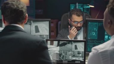 Profesyonel siber güvenlik görevlilerinden oluşan çeşitli erkek takımının omuz üstü çekimleri bilgisayar ekranlarındaki CCTV kameralarını izliyor ve geceleri karanlık ofiste çalışırken telsizle konuşuyor.