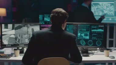 Çoklu bilgisayarın önünde oturan erkek siber güvenlik memurunun Dolly-out arka plan görüntüsü CCTV kameralarını ve ağ veri bilgilerini gece ofiste izlerken görüntüler.